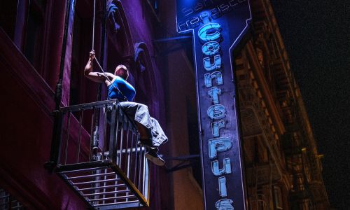 A person with dark skin wearing a blue shirt and gray pants sits on the edge of a large metal cage suspended from the side of a building, hanging from a wire. A blue neon sign reads “San Francisco CounterPulse”