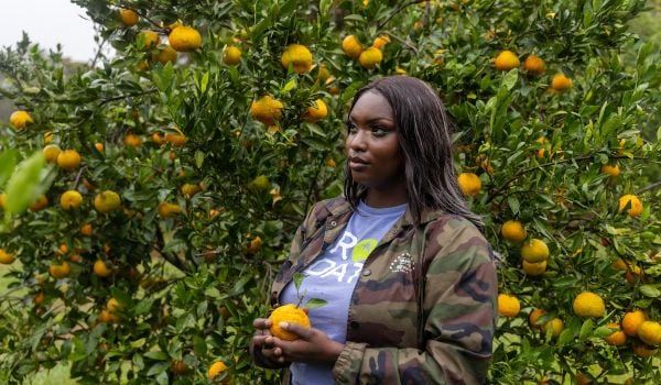 Jonshell Johnson-Whitten holds a satsuma, a type of orange, and stands in front of a tree with more of them