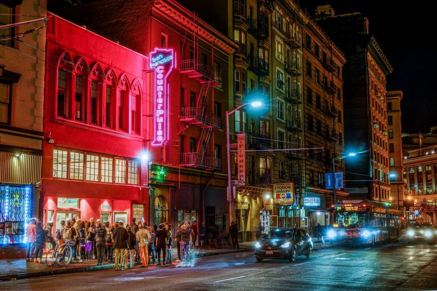 Alt text: A crowd gathers on the street outside a building at night, illuminated by a neon pink sign that reads “San Francisco CounterPulse”