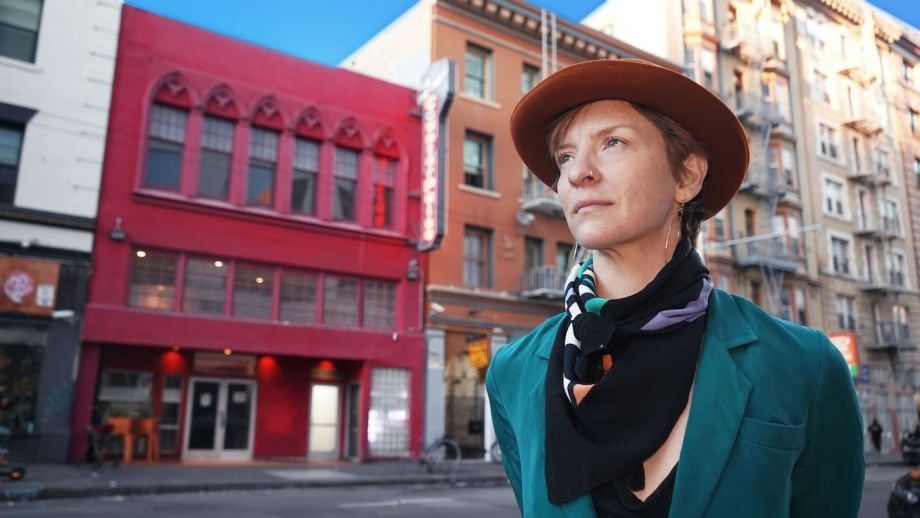 Alt text: Julie Phelps, a person with light skin wearing a green jacket, black patterned scarf, and a brown fedora, stands in front of the magenta facade of CounterPulse’s building.