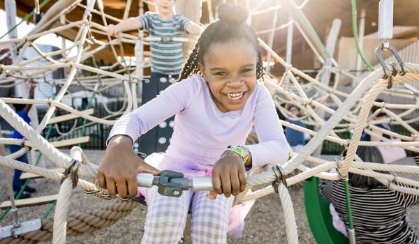 One child smiles while playing on a rope fixture on a playground; another child can be seen in the background