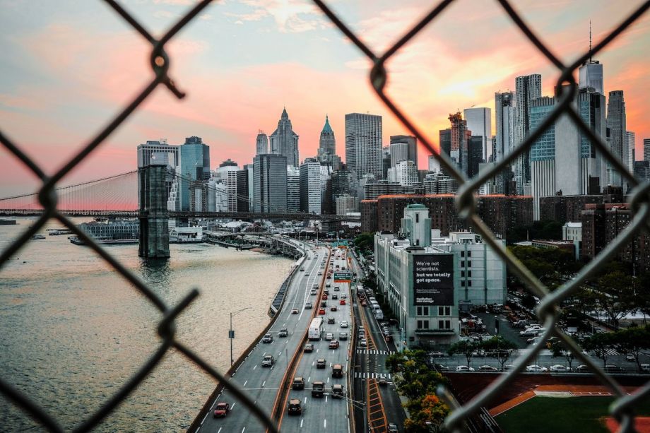 Looking through a gate toward lower Manhattan in New York City. Cars driving on the highway and skyscrapers.