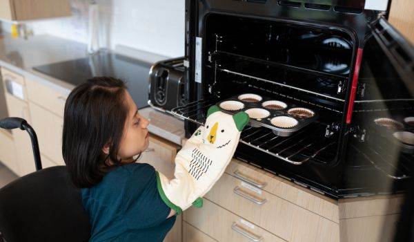 A South Asian person sits in her wheelchair and places a muffin pan into a mid-height oven with side-hinged door.