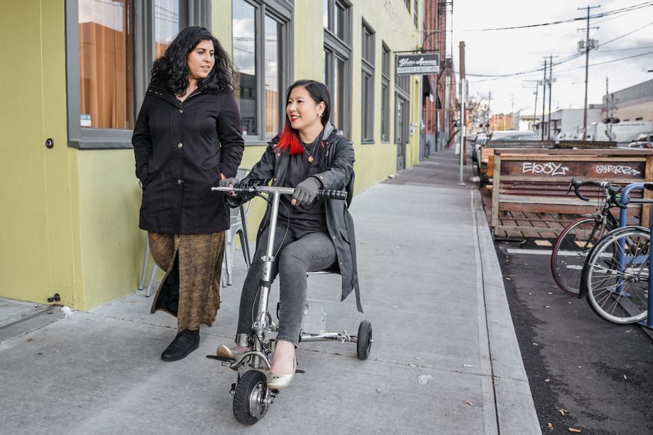 A Latinx (invisibly) disabled woman talking and walking alongside her friend, an Asian disabled genderfluid person wearing compression gloves and driving a lightweight electric mobility scooter.
