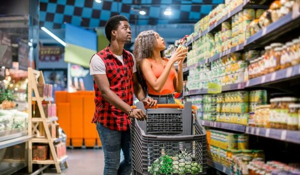 Two people look at a shelf on an aisle of a grocery store; a shopping basket is in front of them.