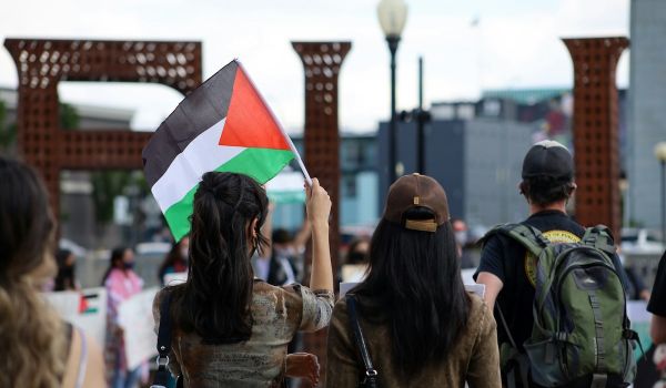 The backs of three people; the person on the left holds up a Palestinian flag.