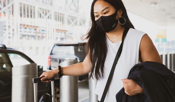 Person wears black mask over face and a white shirt, while walking with luggage