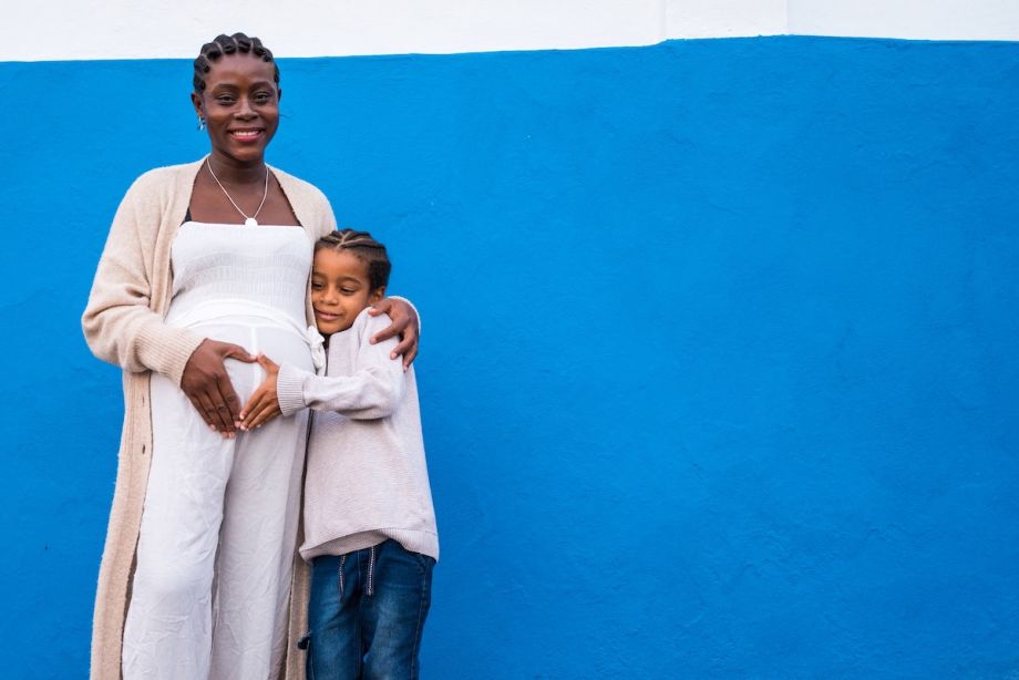 Pregnant person stands in front of a blue wall, with a child hugging their left side